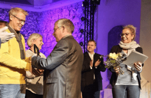 Preisverleihung - 5 Personen stehen auf der Bühne vor einem lila angeleuchteten Hintergrund. Herr Schwarze gratuliert Herrn Schimmel vom Bündnis, die anderen applaudieren. Frau Schade hält die Urkunde und einen Strauß Blumen in ihren Händen.