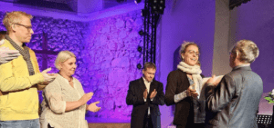 Preisverleihung - 5 Personen stehen auf der Bühne vor einem lila angeleuchteten Hintergrund. Herr Schwarze gratuliert Frau Schade vom Bündnis, die anderen applaudieren.