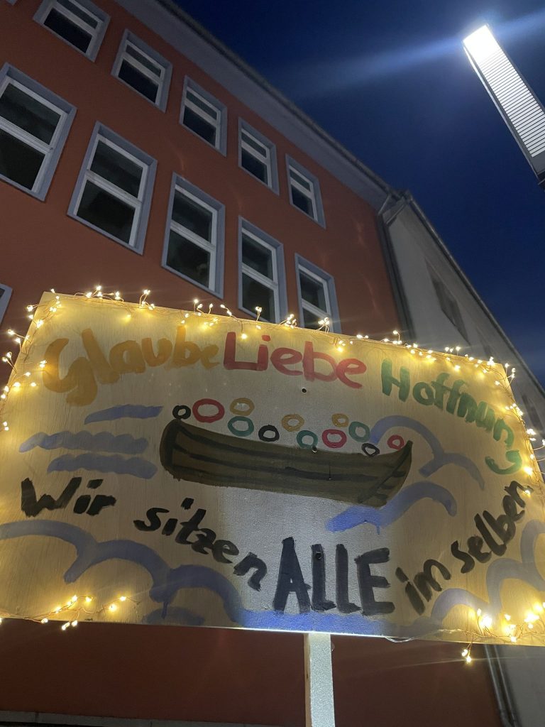 Ein Plakat mit einem gemalten Boot, in dem Menschen sitzen. Text: Glaube, Liebe Hoffnung - Wir sitzen alle im selben Boot