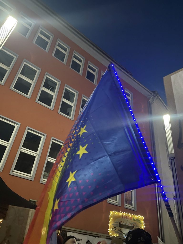 Eine blau beleuchtete Regenbogen-Europa-Flagge, im Hintergrund ein oranges Gebäude